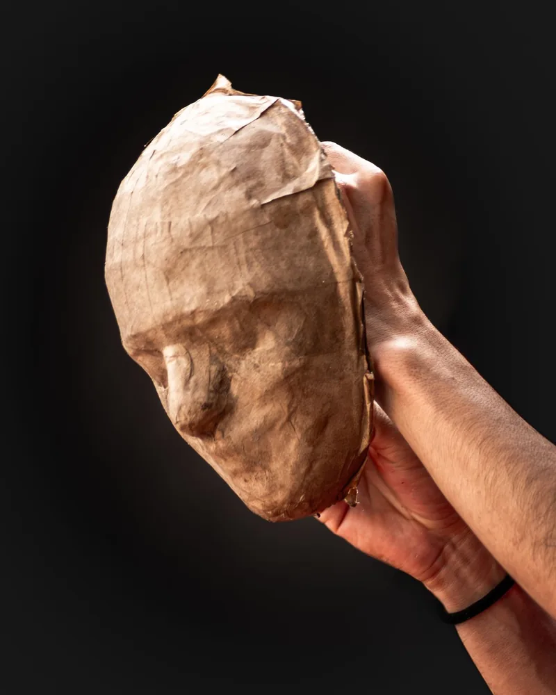 Máscara de papel encolado sin pintar sostenida en la mano sobre fondo oscuro
